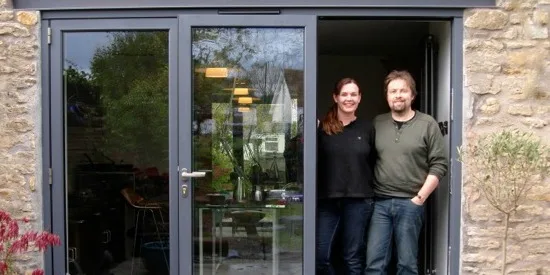 A couple standing in the open bifold doorway of their stone-built home